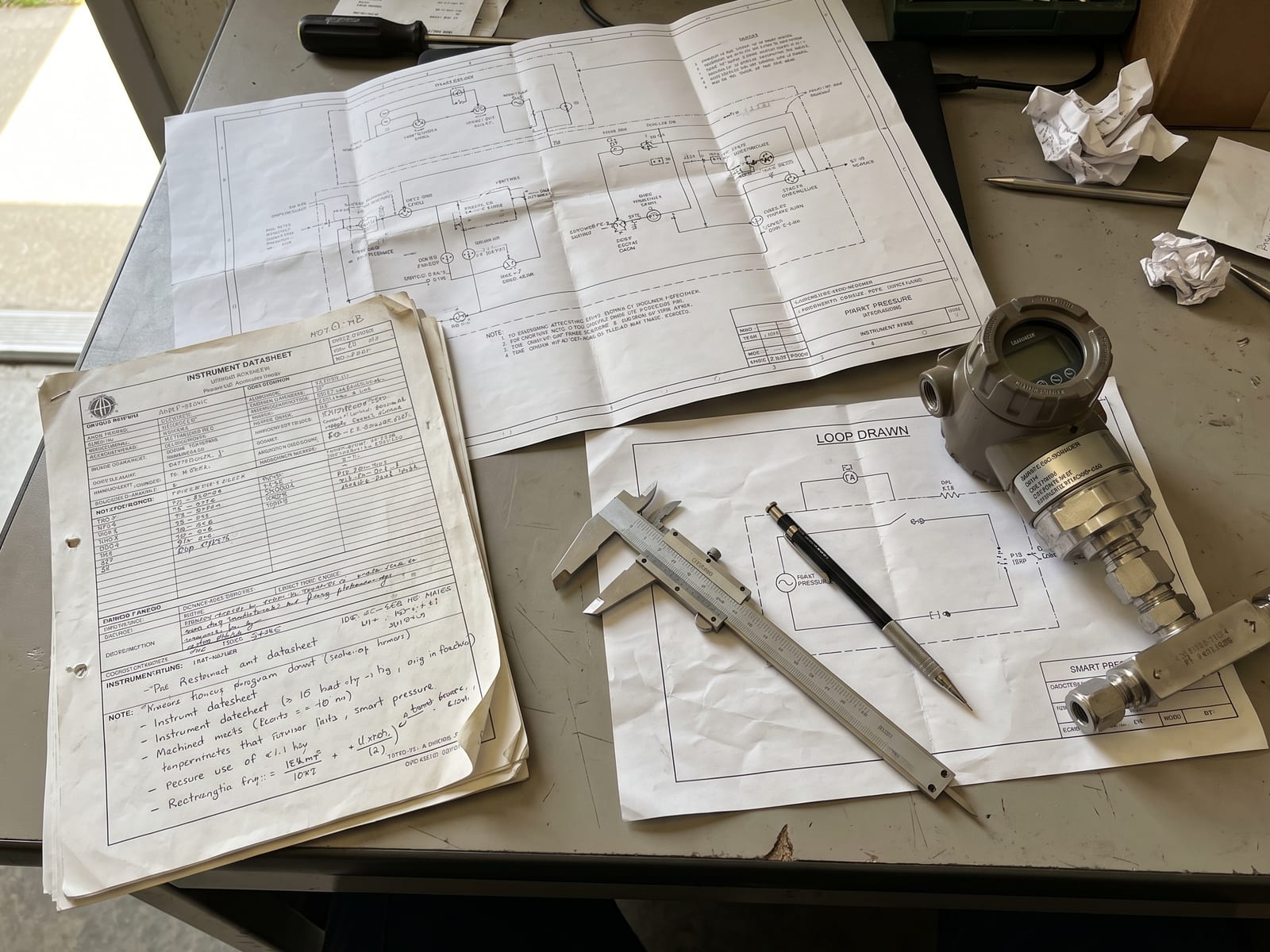 An instrument datasheet, a marked-up loop drawing and a set of calipers laid out on a desk beside a pressure transmitter.